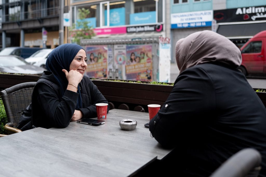 Esra Karamik sitzt draußen an einem Bistrotisch neben einer leicht befahrenen Straße. Auf der anderen Straßenseite reihen sich Läden aneinander. Esra Karamik ist links im Bild und blickt mit einem offenen Lächeln in Richtung einer zweiten Person rechts im Bild an der gegenüberliegenden Seite des Tisches. Beide haben ihre Ellenbogen auf dem Tisch liegen und sind zueinander gewandt. Esra Karamik stützt ihren Kopf auf ihre rechte Hand, am Handgelenk trägt sie einen goldenen Armreif. Sie trägt ein Nasenpiercing, ihre Augen sind leicht geschminkt. Beide Frauen tragen schwarze, langärmelige Oberteile und einen Hijab in dunkelblau bzw. hellbraun. Vor ihnen steht jeweils ein Kaffeebecher, daneben liegen ihre Mobiltelefone.