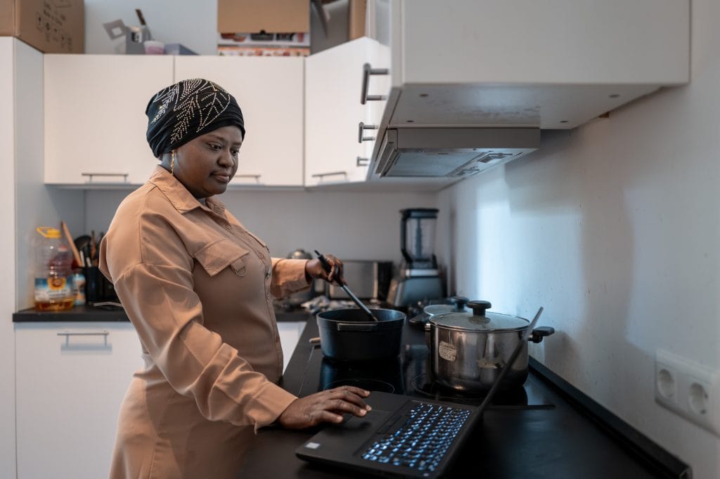 Rofiat Akinremi steht in einer Küche mit weißen Fronten und schwarzer Arbeitsplatte. Ihre rechte Seite ist zur Kamera gedreht, vor ihr, rechts im Bild, stehen drei Töpfe auf dem Herd. In einem rührt sie mit der linken Hand mit einem Kochlöffel. Mit der rechten Hand bedient sie das Mousepad eines Laptops, der neben dem Herd auf der Arbeitsplatte steht. Ihr Blick geht in Richtung des Bildschirms. Rofiat Akinremi trägt ein helles, lachsfarbenes Hemdkleid mit langen Ärmeln und einen dünnen schwarzen Turban, der mit Blättern aus Pailletten  geschmückt ist. Sie trägt einen goldenen Ohrring. Im Hintergrund stehen Küchenutensilien auf der Arbeitsfläche, auf den Hängeschränken sind einige Kartons zu sehen.