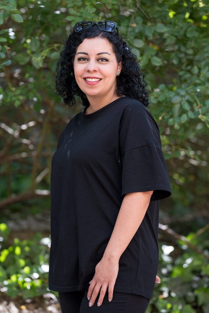 Ebru Schaefer steht draußen vor einem Strauch mit grünen Blättern, auf den Sonnenlicht fällt. Ihre linke Schulter ist zur Kamera gedreht, sie blickt mit einem offenen Lächeln in die Kamera. Sie trägt ein schwarzes, weites, kurzärmliges Tshirt und eine schwarze Hose, ihre Hand liegt auf ihrem linken Bein auf. Sie trägt Lippenstift und Lidschatten, ihre Fingernägel sind lackiert. Ihre lockigen schwarzen Haare werden von einer schwarzen Sonnenbrille nach hinten gehalten.