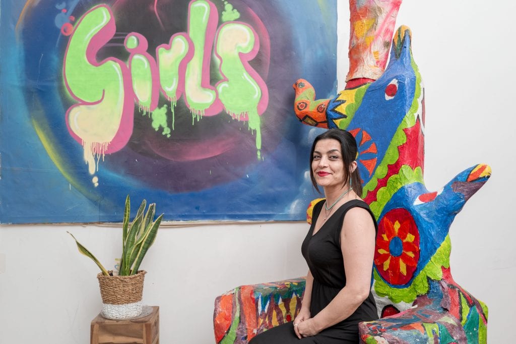 Eylem Bozkaya sitzt aufrecht auf einem sehr großen Stuhl, der eher einer Skulptur in fantasievollen Formen gleicht. Die Skulptur ist aus Pappmache und mit bunten Mustern bemalt. Im Hintergrund hängt an einer weißen Wand eine große Leinwand, die blau und lila  bemalt ist. In Pink, grün und weiß ist das Wort “Girls” aufgesprüht. Eylem Bozkaya blickt mit einem Lächeln in die Kamera. Ihre dunkelbraunen, glatten Harre trägt sie in einem Zopf, links und rechts umspielen Strähnen ihr Gesicht. Ihre Augen sind leicht geschminkt und sie trägt roten Lippenstift. Sie trägt einen türkisen Ohrring und zwei Ketten, eine davon aus türkisenen Steinen. Sie trägt ein schwarzes, ärmelloses Kleid. Vor ihr steht auf einem Holzblock eine Zimmerpflanze.