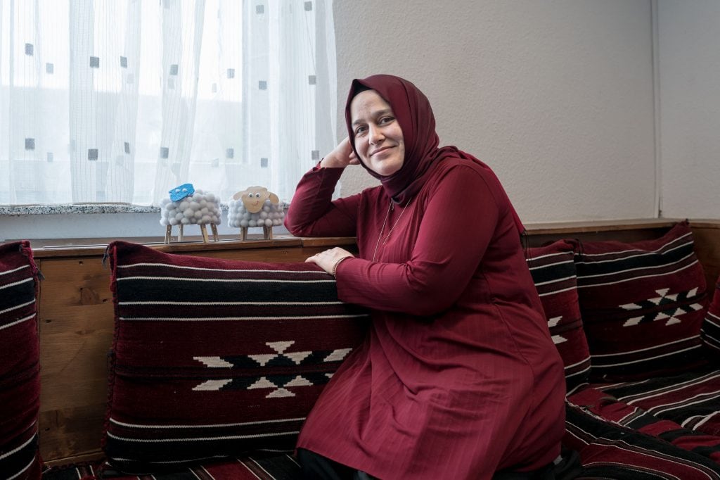 Sennur Tufan kniet auf einer breiten Holzbank und lehnt sich an ein großes Kissen aus grob gewebtem Stoff. Mit demselben Stoff ist auch die Sitzfläche bezogen. Ihr rechter Arm liegt auf der Lehne der Bank, sie stützt ihren Kopf auf der rechten Hand ab, ihr Blick geht in Richtung Kamera, sie lächelt leicht. Sie trägt einen Hijab in weinrot und ein knielanges Oberteil in ähnlicher Farbe sowie eine schwarze Hose. Auf der Lehne der Bank stehen zwei kleine gebastelte Schafe aus vielen weißen Wattebällchen, je vier Beinen aus Holzwäscheklammern, einem Gesicht aus Papier und aufgeklebten Wackelaugen. Licht fällt durch ein Fenster, das von einer hellen Gardine verdeckt ist