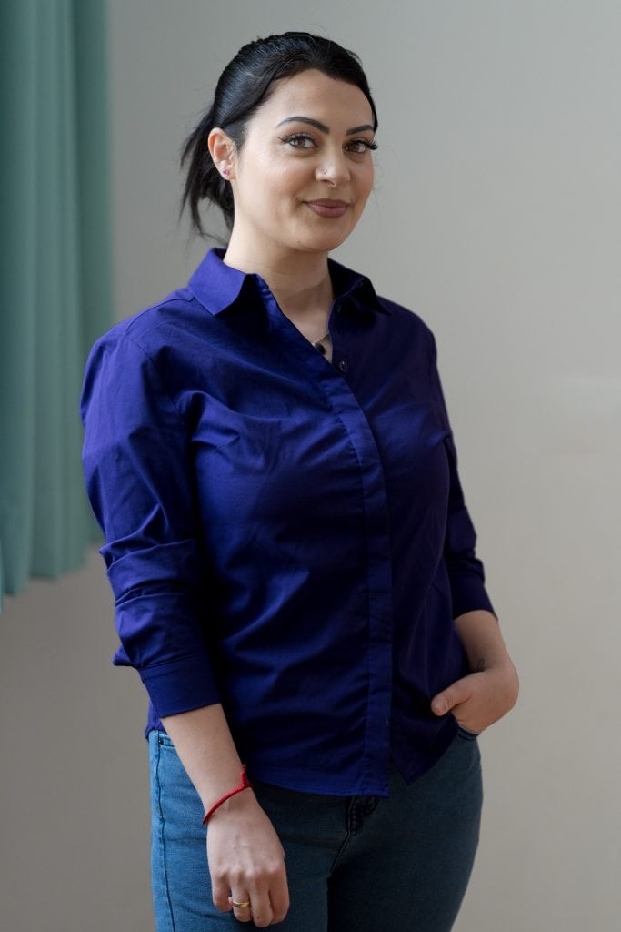 Gardine erkennbar. Sie blickt mit einem Lächeln in die Kamera, ihre dunkelbraunen, glatten Haare hat sie zu einem Zopf nach hinten gebunden. Sie trägt eine Jeans und eine blaue langärmelige Bluse, die Ärmel sind etwas nach oben gekrempelt. Um den Hals trägt sie eine Kette, und an ihrem rechten Handgelenk einen Anhänger an einem roten Band. An ihrem rechten Ringfinger trägt sie einen goldenen Ring. Die linke Hand steckt in der linken vorderen  Hosentasche. Ihre Augen sind geschminkt, sie trägt Lippenstift und ein Nasenpiercing. An ihrem rechten Ohr trägt sie einen Ohrring in Form eines kleinen violetten Herzen.