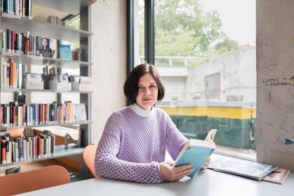 Nadjeschda Pugatschowa sitzt an einem Tisch mit weißer Oberfläche in einem Raum vor einem großen Fenster. Links und rechts des Fensters ist eine unverputzte Betonwand zu erkennen, verschiedene Menschen haben Namen und Herzen auf die Wand gemalt. Auch im Bildhintergrund steht ein schlichtes Bücherregal, die Bücher sind sortiert und tragen Kennzeichnungen einer Bibliothek. Nadjeschda Pugatschowa hält ein aufgeschlagenes Buch in beiden Händen, ihre Unterarme liegen auf dem Tisch ab. Sie blickt in die Kamera. Ihre glatten, dunkelbraunen Haare trägt sie in einem Kurzhaarschnitt bis zum Kinn, unter ihrem langärmligen, fliederfarbenen Pullover trägt sie ein weißes Rollkragenshirt