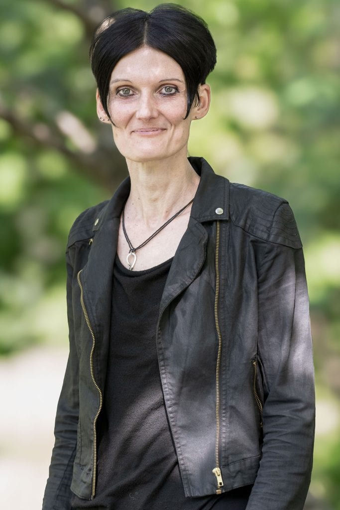 Jeanine Fornaçon steht draußen vor einem sonnigen Hintergrund, der Bäume und eine Wiese vermuten lässt. Sie blickt mit einem Lächeln in die Kamera, ihre rechte Schulter ist etwas in den Bildhintergund gedreht. Ihre glatten schwarzen Haare trägt sie in einem Kurzhaarschnitt, links und rechts umrahmt eine Strähne ihr Gesicht. Ihre Augen sind geschminkt, rechts trägt sie drei kleine Ohrringe, links einen. Über einem schwarzen Tshirt trägt sie offen eine Bikerjacke aus schwarzem Stoff. Um ihren Hals trägt sie an einem dünnen Lederband einen silbernen Anhänger.