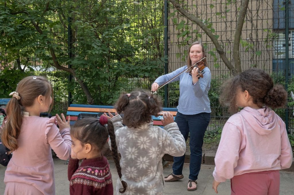 Miriam Stute steht draußen auf quadratischen Betonplatten vor einem hohen Drahtzaun, an dem Pflanzen hochwachsen. Dahinter ist eine Hauswand zu erkennen. Neben ihr steht eine in verschiedenen Farben gestrichene Bank. Sie trägt ein langärmliges, hellblaues T Shirt, eine dunkelblaue Hose und Sandalen. Ihre Nägel sind rotlackiert. Ihre schulterlangen, glatten blonden Haare trägt sie offen. Auf ihrer linken Schulter liegt eine Geige auf, mit der linken Hand hält sie die Geige, ihre Finger liegen auf den Saiten. In der rechten Hand hält sie einen Geigenbogen, der am anderen Ende quer zu den Saiten auf der Geige aufliegt. Sie blickt in den Bildvordergrund, in dem vier Kinder im Grundschulalter mit ihren Rücken zur Kamera in ihre Richtung schauen. Die Hände der Kinder sind in Bewegung, als würden sie winken oder klatschen.
